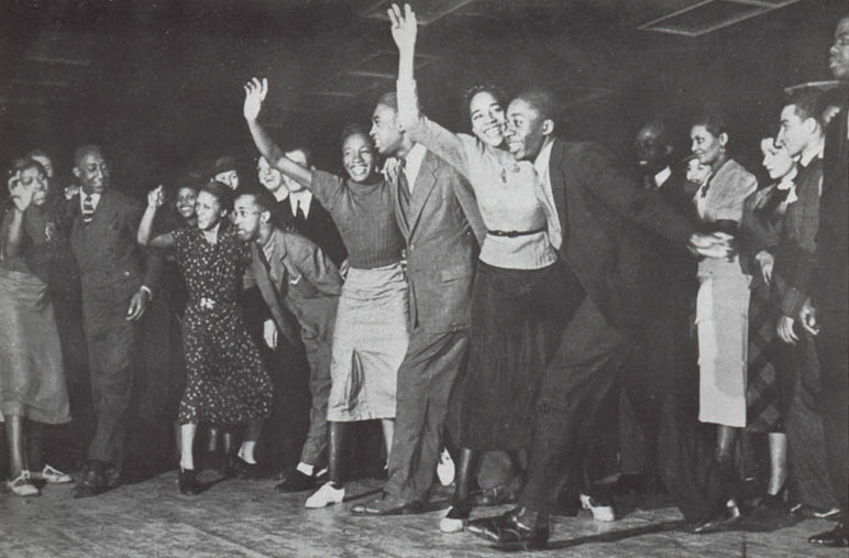 Black-and-white photograph of a lively line of swing dancers at the edge of the Savoy Ballroom floor, several couples mid-step with arms raised and faces beaming, surrounded by a crowd of cheering onlookers.