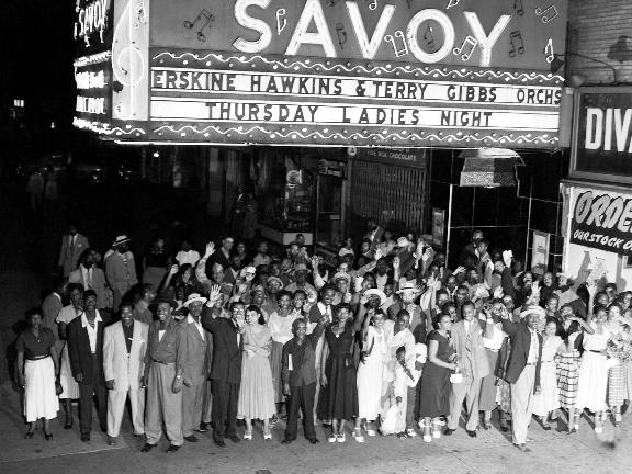 Black-and-white night photograph of a large crowd posing and waving beneath the illuminated marquee of Harlem's Savoy Ballroom, the sign advertising the Erskine Hawkins and Terry Gibbs orchestras on Thursday Ladies Night.