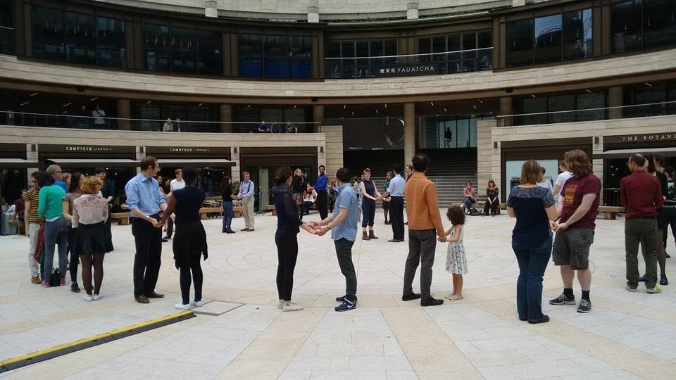 Dance students standing in a circle holding hands during an outdoor swing dance class, lesson in Broadgate Circle (London) with Kibble and Robyn teaching from the center of the circle