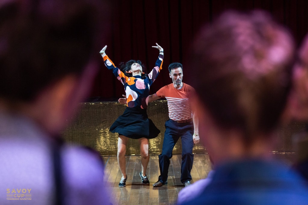 Colour photograph of two swing dancers on a softly lit stage, the woman (Michaela) with both arms raised in a dramatic pose beside her partner (Kibble) seen past the blurred silhouettes of the audience, at the Savoy Cup festival.