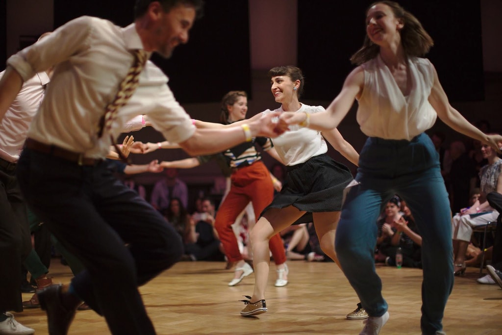 Dancers in vintage-style clothing swing dancing energetically at a social dance event with Kibble and Judith in the foreground