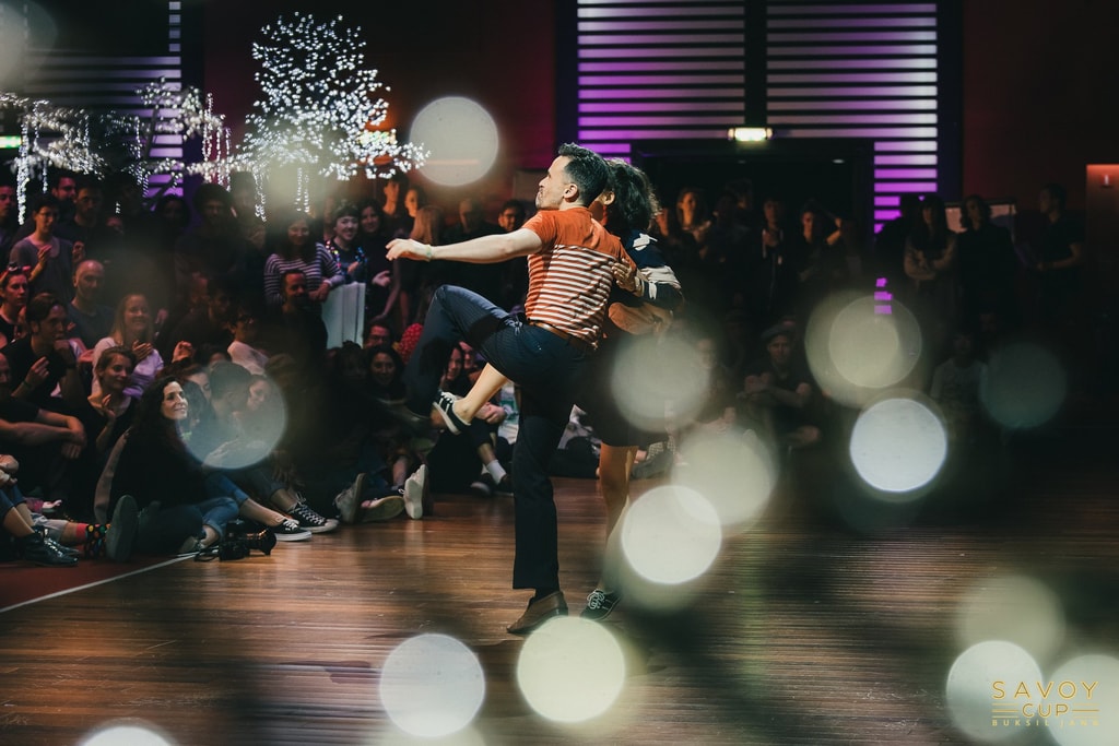 Colour photograph of a couple performing an energetic swing dance routine on stage in front of a seated audience, with sparkling fairy lights glowing in the background, at the Savoy Cup festival.